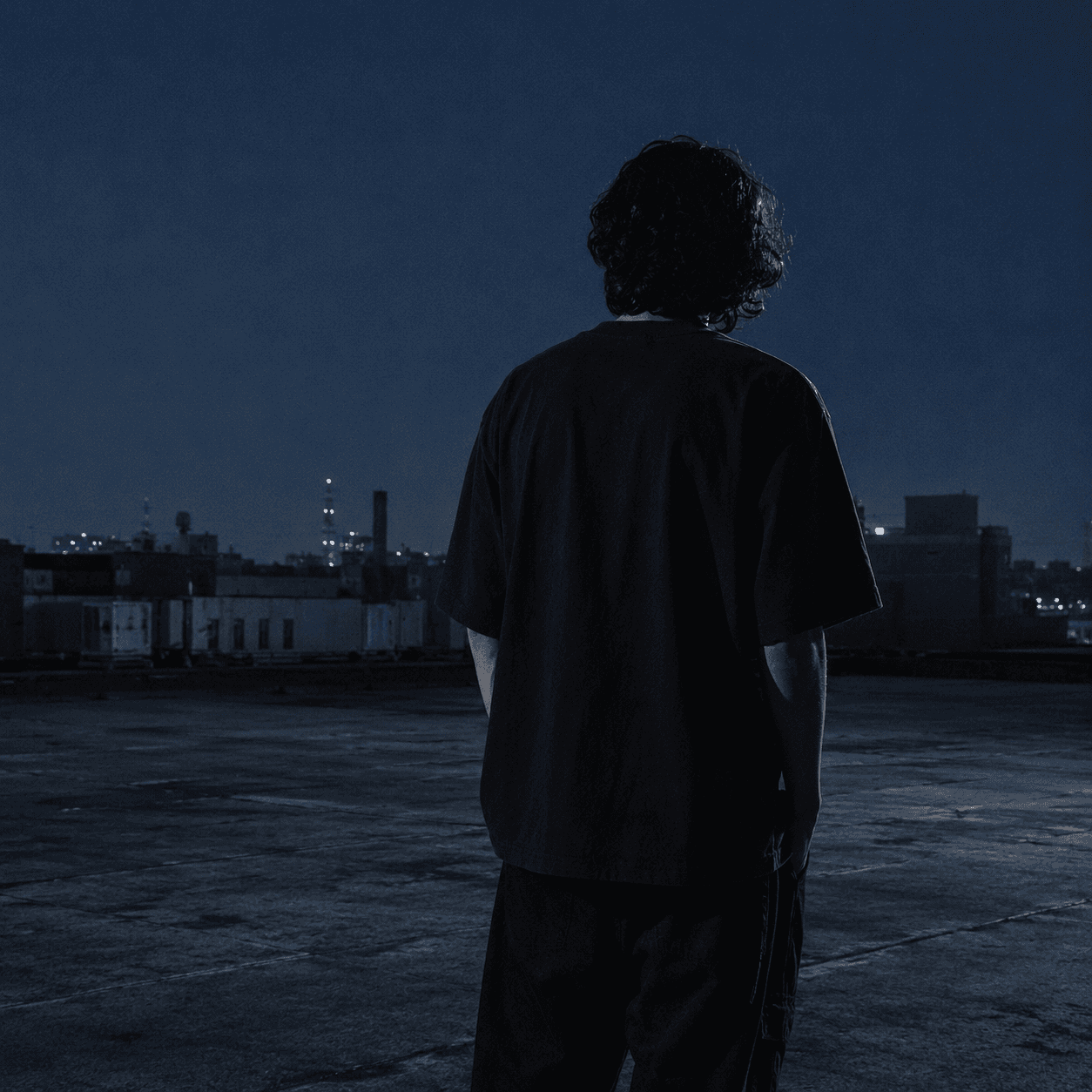 Figure in the Mr. Exotixx tee on a California rooftop at blue hour, looking out over a distant industrial skyline