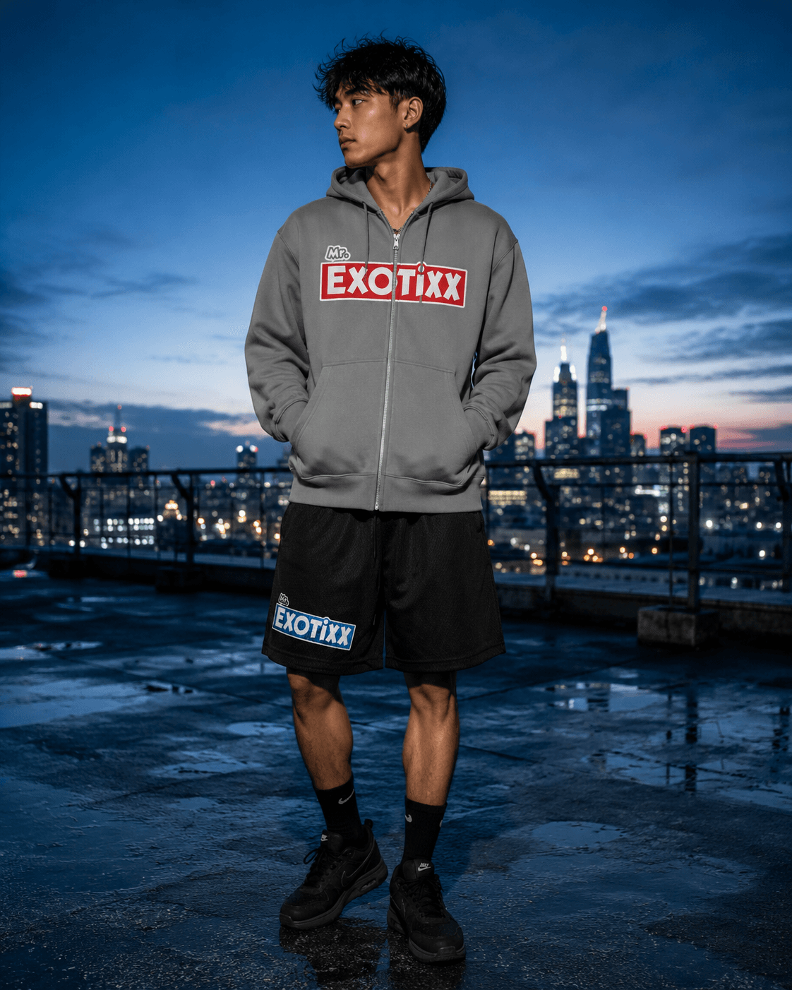 Model in gray Mr. Exotixx zip-up hoodie and branded shorts on a rooftop at dusk with city skyline behind